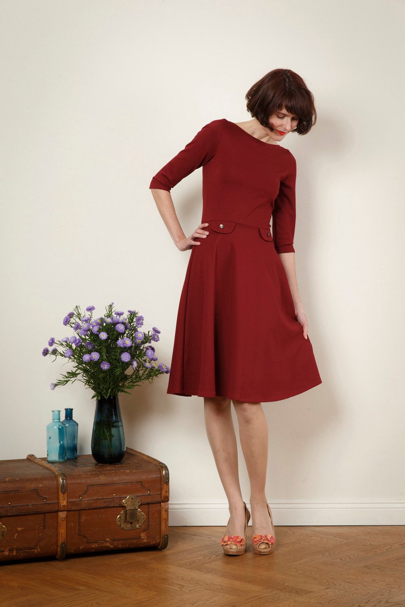 May include: A woman wearing a maroon dress with a fitted bodice and a full skirt. She is standing on a wooden floor and has a bouquet of purple flowers in a vase on a wooden chest behind her.