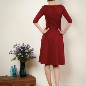 May include: A woman wearing a maroon dress with a scoop neck and three-quarter sleeves. She is standing in front of a white wall with her hands on her hips.