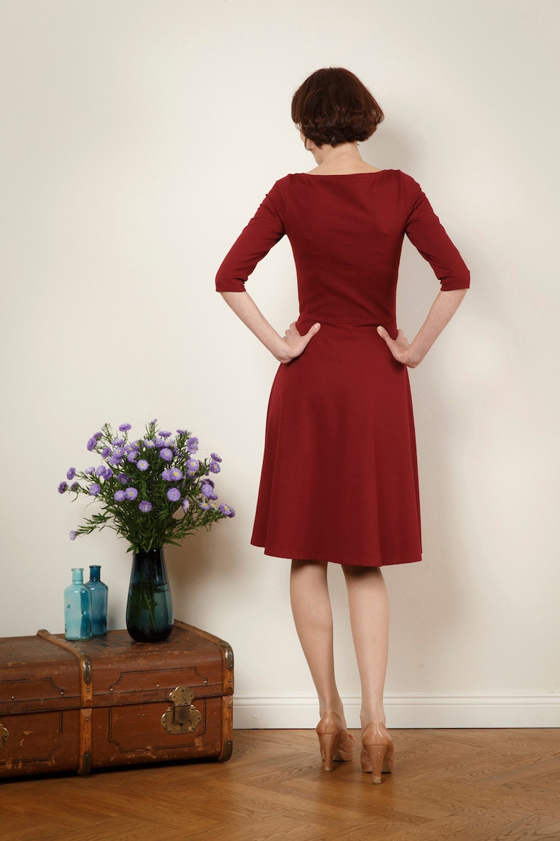 May include: A woman wearing a maroon dress with a scoop neck and three-quarter sleeves. She is standing in front of a white wall with her hands on her hips.