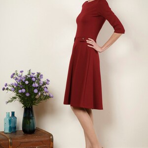 May include: A woman wearing a maroon dress with a fitted bodice and a flared skirt. She is wearing nude heels with floral embellishments. The woman is standing in front of a white wall with a wooden floor.