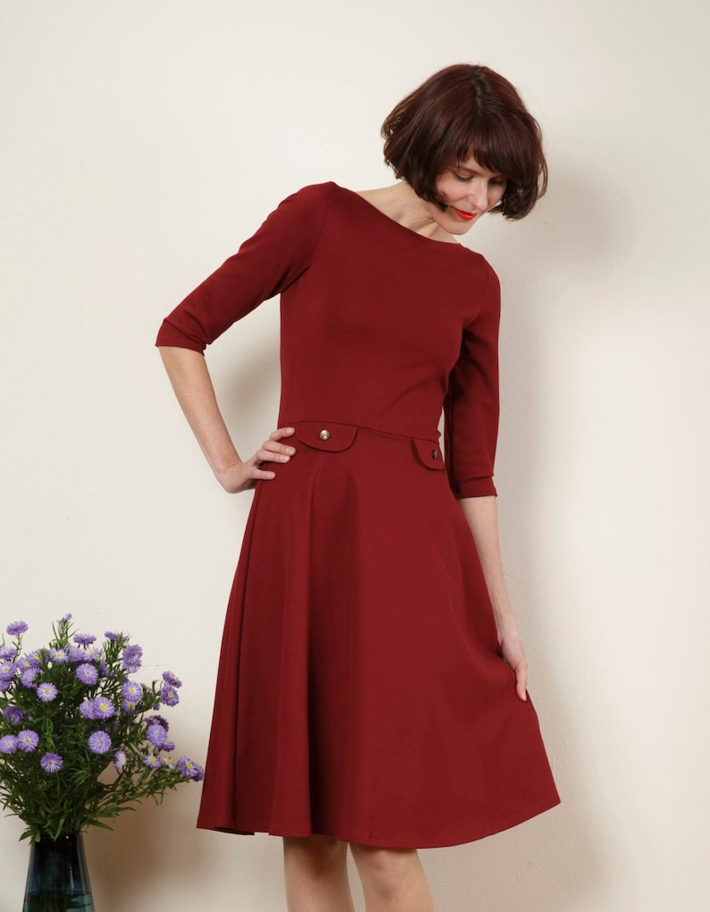 May include: A woman wearing a maroon dress with a boat neck and three-quarter sleeves. The dress has a fitted bodice and a flared skirt.