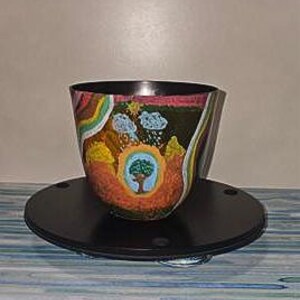 May include: A dark-coloured bowl featuring a hand-painted landscape design. The artwork depicts a central tree surrounded by an orange aura, with blue clouds, rain, a yellow sun, and rainbow stripes on the sides. The bowl sits on a black circular base.