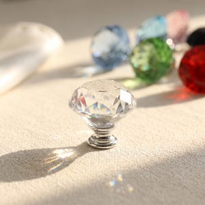 May include: A clear, diamond-shaped glass cabinet knob with a silver-toned metal base. The faceted surface reflects light, casting a prismatic shadow. In the blurred background, several other knobs in various colours are visible, including blue, green, and red.