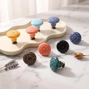 Puede incluir: Una colección de pomos redondos de cerámica para armarios en varios colores, incluyendo amarillo, azul claro, azul oscuro, coral, negro, marrón y verde azulado. Cada pomo presenta un patrón floral en relieve y una base metálica.