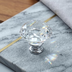May include: A clear, faceted, diamond-shaped glass knob with a polished chrome metal base. The knob features multiple triangular facets that catch the light, creating subtle rainbow refractions on its surface and the surrounding area.