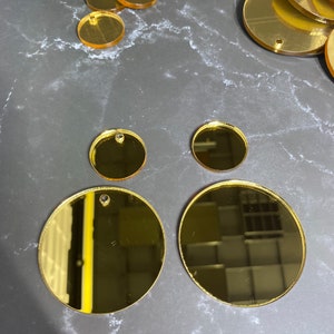 May include: Various sizes of gold-coloured, reflective acrylic circles, some with small holes for hanging. The circles are arranged on a dark grey, marbled surface. Suitable for crafting or jewellery making.