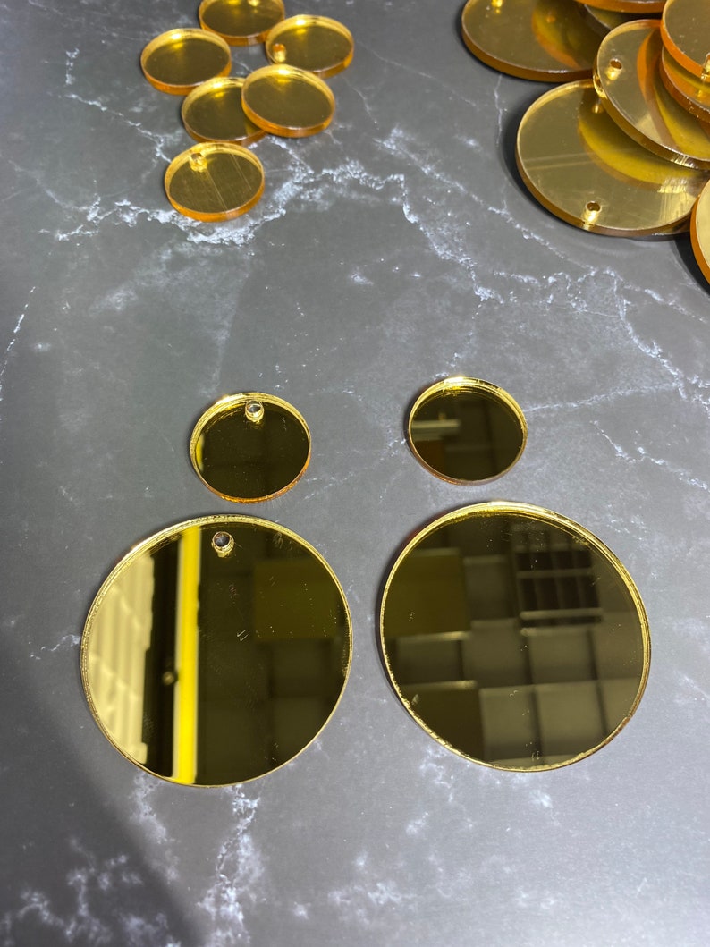 May include: Various sizes of gold-coloured, reflective acrylic circles, some with small holes for hanging. The circles are arranged on a dark grey, marbled surface. Suitable for crafting or jewellery making.