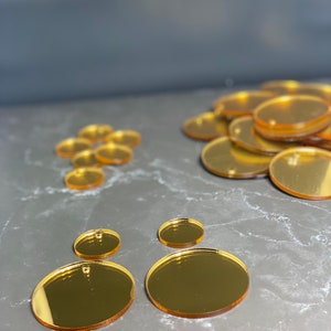 May include: A selection of gold-coloured, oval-shaped acrylic blanks. The blanks are of varying sizes, some with a small hole at the top for hanging. The reflective surface of the blanks creates a mirror-like effect. Suitable for craft projects.
