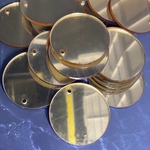 May include: A pile of gold-coloured, circular acrylic blanks with small holes near the edge. The blanks are reflective and appear to be stacked on a dark, marbled surface. Suitable for crafting or jewellery making.