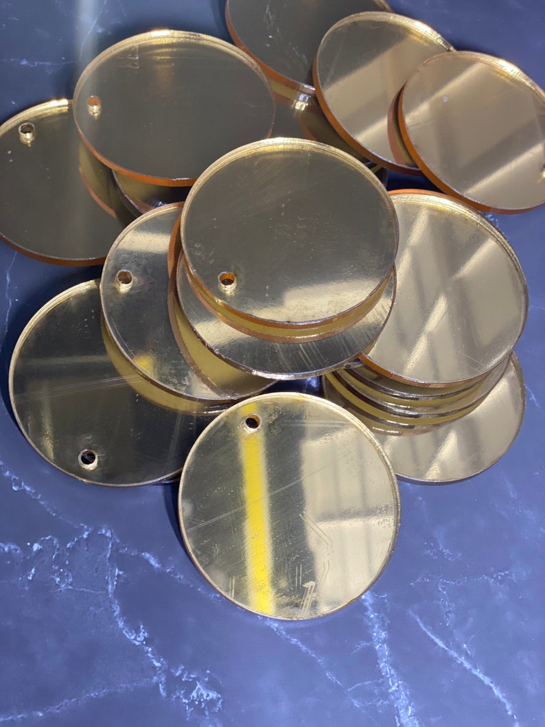 May include: A pile of gold-coloured, circular acrylic blanks with small holes near the edge. The blanks are reflective and appear to be stacked on a dark, marbled surface. Suitable for crafting or jewellery making.