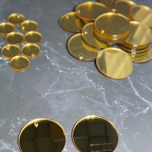 May include: A selection of gold-coloured, circular acrylic blanks with small holes for crafting. The blanks are arranged in several groups on a grey surface. The reflective surface of the blanks creates a shiny appearance.