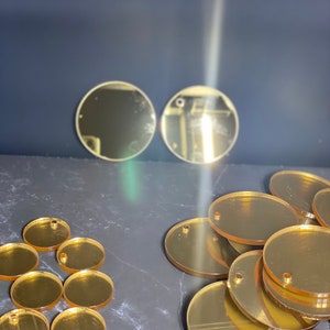 May include: Various gold-coloured acrylic circles and ovals, some with pre-drilled holes. The shapes are arranged on a dark surface, with a few circles placed separately in the background. Suitable for crafting or jewellery making.