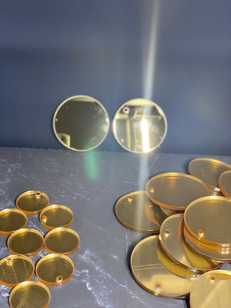 May include: Various gold-coloured acrylic circles and ovals, some with pre-drilled holes. The shapes are arranged on a dark surface, with a few circles placed separately in the background. Suitable for crafting or jewellery making.