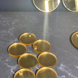 May include: Several gold-coloured, circular acrylic blanks with pre-drilled holes, suitable for jewellery making or crafts. The reflective blanks are arranged on a dark surface, with a few larger circles visible.