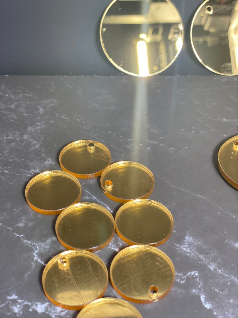 May include: Several gold-coloured, circular acrylic blanks with pre-drilled holes, suitable for jewellery making or crafts. The reflective blanks are arranged on a dark surface, with a few larger circles visible.