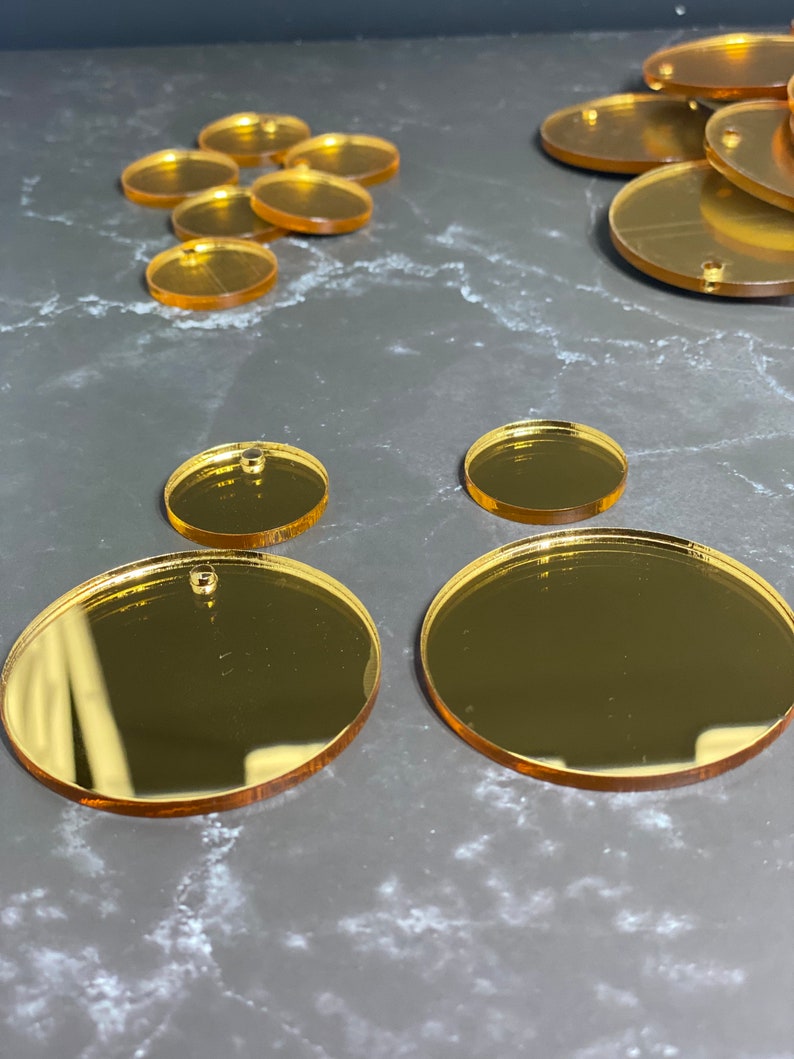 May include: Various gold-coloured acrylic blanks in oval and circular shapes, ideal for crafting. The blanks have small holes and come in different sizes, with larger ovals and smaller circles. The reflective surface gives them a shiny appearance.