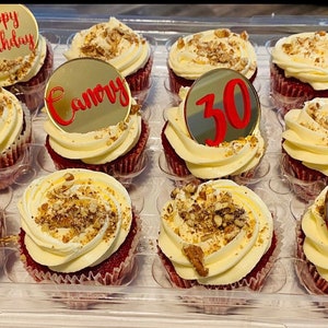 May include: A tray of twelve red velvet cupcakes, each topped with white frosting and chopped nuts. Some cupcakes have gold toppers with red text saying "Happy Birthday", "Camory", and the number "30".