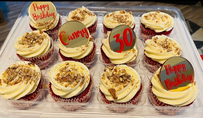 May include: A tray of twelve red velvet cupcakes, each topped with white frosting and chopped nuts. Some cupcakes have gold toppers with red text saying "Happy Birthday", "Camory", and the number "30".