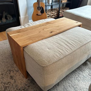Peut inclure: Un plateau rectangulaire en bois avec une finition naturelle en grain de bois brun clair, conçu pour reposer sur le bras ou l'assise d'un grand pouf beige rembourré. Le plateau présente une surface plane et deux panneaux latéraux verticaux.