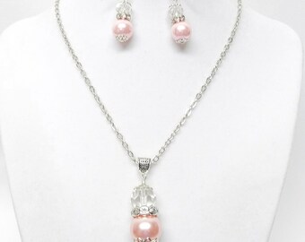 Puede incluir: Un collar de cadena de plata con un colgante de perla rosa y pendientes a juego. El colgante presenta una cuenta de cristal transparente sobre la perla. Los pendientes tienen una cuenta de cristal transparente sobre cada perla rosa.