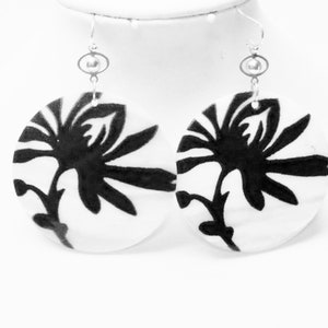 May include: A pair of white, circular earrings with a black floral design. The earrings have silver-colored hooks and small, oval-shaped accents. The black floral pattern is a stylized depiction of a flower.