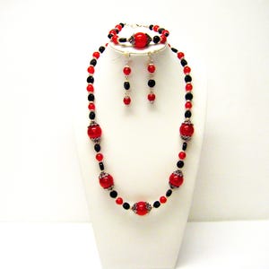 Op de afbeelding: Een rode en zwarte kralenset met ketting, armband en oorbellen. De ketting en armband hebben grote rode kralen met zilveren accenten. De oorbellen zijn zilver met rode kralen.