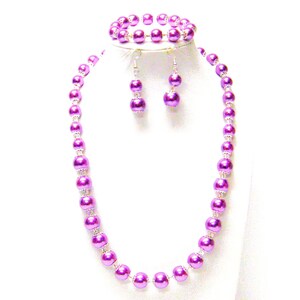 Puede incluir: Un conjunto de joyería de tres piezas que incluye un collar, una pulsera y pendientes. El conjunto presenta grandes cuentas redondas de color púrpura brillante con espaciadores plateados. Los pendientes tienen un diseño colgante.