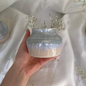 Könnte beinhalten: Eine kleine Keramikvase mit bauchigem Körper und engem Hals. Der obere Teil ist mit einer glänzenden, irisierenden hellblauen Glasur überzogen, die über den unteren Teil läuft, welcher eine glatte, matte beigefarbene Tontextur aufweist.