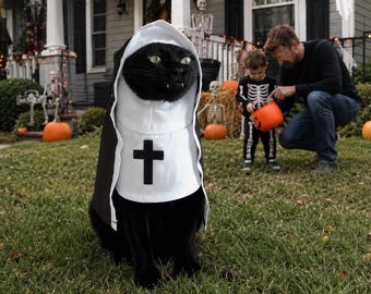 May include: A black cat wearing a white and black habit costume with a hood and a black cross emblem on the chest. The costume covers the head and torso of the cat, featuring a white face frame and a black cape-like back section with white trim.