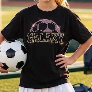 May include: A black t-shirt featuring a rhinestone soccer ball graphic above the word GALAXY in gold rhinestones, paired with a white pleated skirt and a black baseball cap with a matching rhinestone soccer ball and GALAXY text design.