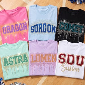 Op de afbeelding: Zes T-shirts met korte mouwen in roze, lichtblauw, zwart, mintgroen, lavendel en wit. Elk shirt heeft een uniek woord in strass-letters: ORAGON, SURGON, COMET, ASTRA, LUMEN en SDU Susion, met decoratieve druppelvormige accenten.