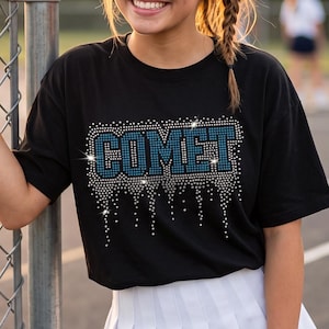 Puede incluir: Una gorra de béisbol negra con una letra C azul, una camiseta negra con la palabra COMET en pedrería azul y plateada con efecto de goteo, y una falda plisada blanca con una franja azul a lo largo del dobladillo inferior de la prenda.