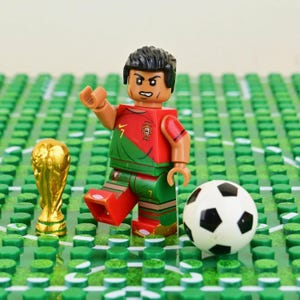 May include: A plastic minifigure wearing a red and green soccer jersey with the number 7 and a team crest, standing next to a gold-colored trophy and a black and white soccer ball on a green studded baseplate representing a soccer field.