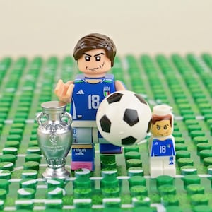 Pode incluir: Uma minifigura de plástico vestindo uma camisa de futebol azul da Itália com o número 18, segurando uma bola de futebol preta e branca, acompanhada por uma minifigura menor correspondente e uma réplica de troféu prateada sobre uma base verde.