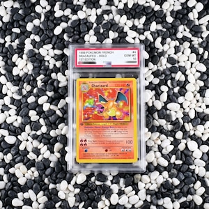May include: A 1999 1st Edition French Charizard holographic trading card graded Gem Mt 10, encased in a clear protective plastic slab. The card features a fire-breathing dragon illustration, 120 HP, and text detailing its Energy Burn power.