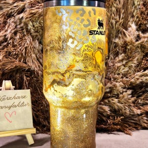 Può includere: Un bicchiere Stanley dorato con finitura glitterata e motivo leopardato sulla parte superiore. Il bicchiere ha un coperchio nero con un dettaglio arancione e una cannuccia. Un piccolo cartello mostra il testo scritto a mano Herzharz Manufaktur con un cuore.