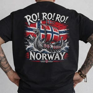 May include: A black t-shirt featuring a graphic on the back. The design displays a Viking longship sailing on waves in front of a Norwegian flag. The text reads RO! RO! RO! at the top and NORWAY at the bottom, with a small Norwegian flag icon below.