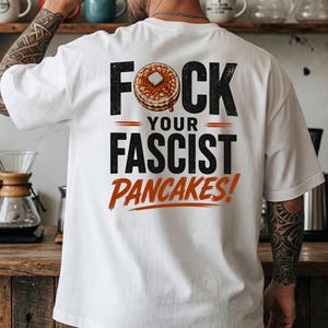 Può includere: Una t-shirt bianca a maniche corte con una grafica sul retro che riporta il testo FOCK YOUR FASCIST PANCAKES! e l'illustrazione di una pila di pancake con un pezzetto di burro. La maglietta è indossata con un cappellino da baseball nero.