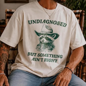 May include: An off-white short-sleeve t-shirt featuring a dark green graphic of a raccoon wearing a cowboy hat. The text above the raccoon reads "UNDIAGNOSED" and the text below reads "BUT SOMETHING AIN'T RIGHT" in a bold, dark green font.