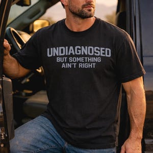 Puede incluir: Una camiseta negra de manga corta y cuello redondo con el texto UNDIAGNOSED BUT SOMETHING AIN'T RIGHT impreso en gris claro en el pecho. La prenda se combina con unos vaqueros azules y una gorra de béisbol de color gris oscuro.