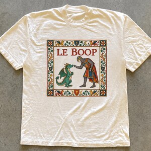 Könnte beinhalten: Ein cremefarbenes Kurzarm-T-Shirt mit einer quadratischen Grafik in der Mitte, die eine mittelalterliche Illustration eines Ritters zeigt, der die Nase eines grünen Drachen berührt. Der Text LE BOOP steht darüber, umrahmt von einer floralen Bordüre.