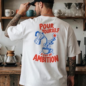 Op de afbeelding: Een wit T-shirt met korte mouwen en een rode en blauwe grafische print op de achterkant. Het ontwerp toont een mokkapot die vloeistof in een koffiekopje schenkt, met een trap die naar een rode ster leidt. De tekst luidt, Pour Yourself A Cup Of Ambition.