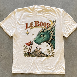 Könnte beinhalten: Ein cremefarbenes Kurzarm-T-Shirt mit einer Grafik eines grünen Drachen, eines Ritters auf einer Leiter und einer Burg. Der Text lautet LE BOOP und THE FORBIDDEN BOOP. Das Design enthält Sterne, florale Ranken und einen roten Umhang.