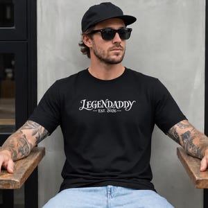 Può includere: Una t-shirt nera a maniche corte con la scritta bianca LEGENDADDY e EST. 2026 stampata sul petto. La maglietta è abbinata a jeans azzurri, un berretto da baseball nero e occhiali da sole neri per un look casual e quotidiano.