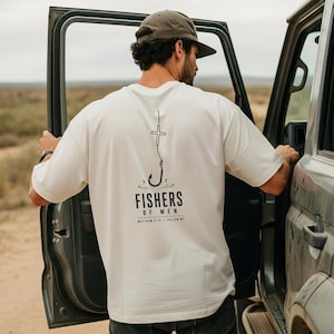 Könnte beinhalten: Ein cremefarbenes Kurzarm-T-Shirt mit einer schwarzen Grafik auf der Rückseite. Das Design zeigt einen Angelhaken mit einem Kreuz an der Spitze, den Text "FISHERS OF MEN" in fetten Buchstaben und den kleineren Text "MATTHEW 4:19 - FOLLOW ME" darunter.