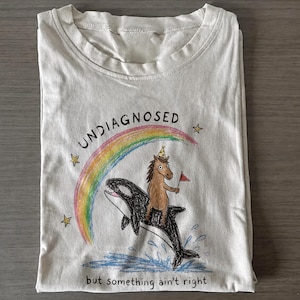 May include: A cream-colored t-shirt featuring a graphic illustration of a brown horse wearing a party hat and holding a red flag while riding a black orca whale. A rainbow arches over the scene with the text UNDIAGNOSED above and but something ain't right below.