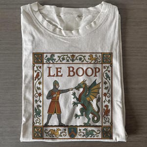 Puede incluir: Una camiseta de color crema con un estampado gráfico de estilo medieval. El diseño muestra a un caballero con cota de malla tocando la nariz de un dragón verde, con el texto LE BOOP centrado encima. La escena está enmarcada por un borde floral.
