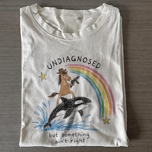 Può includere: Una t-shirt color crema con una grafica in stile disegno a mano di un cavallo marrone che indossa un cappello da cowboy e tiene un'arma da fuoco mentre cavalca un'orca nera. Un arcobaleno, due stelle gialle e spruzzi d'acqua blu circondano le figure. Il testo recita UNDIAGNOSED e but something ain't right.