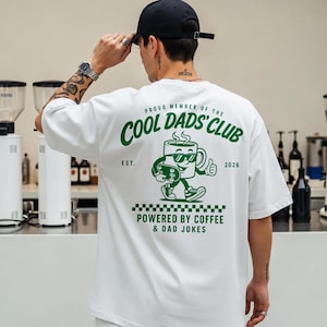 Könnte beinhalten: Ein weißes kurzärmeliges T-Shirt mit einer grünen Grafik auf der Rückseite. Das Design zeigt eine gezeichnete Kaffeetasse mit Sonnenbrille, die ein Skateboard hält, sowie den Text "PROUD MEMBER OF THE COOL DADS' CLUB", "EST. 2026" und "POWERED BY COFFEE & DAD JOKES".