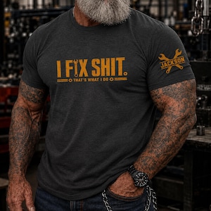 Puede incluir: Una camiseta gris carbón de manga corta con el texto amarillo I FIX SHIT. THAT'S WHAT I DO y un icono de llave inglesa. La manga muestra un gráfico amarillo de dos llaves cruzadas con el nombre JACKSON impreso en el centro del diseño.